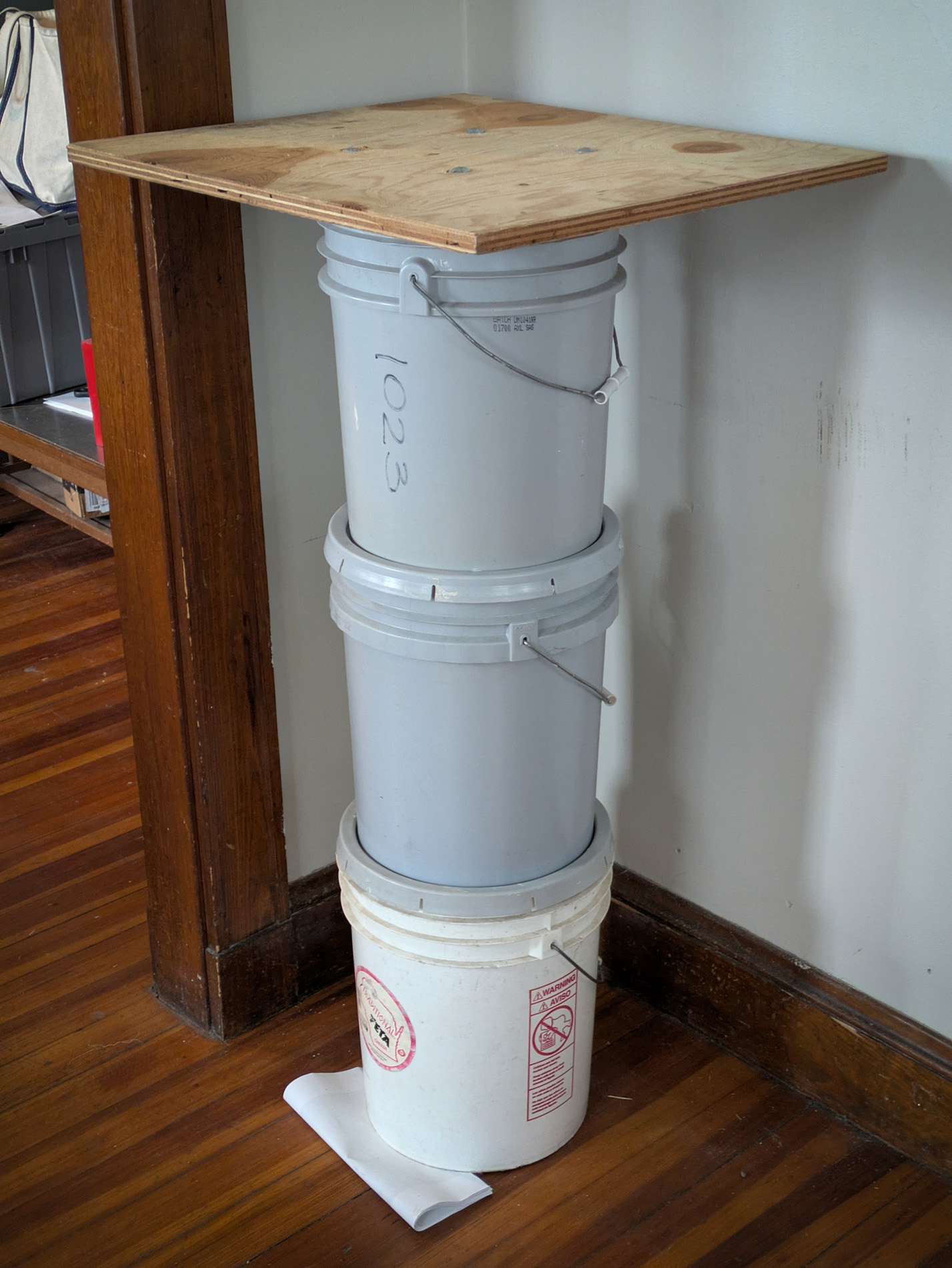 three large PVC buckets stacked with a rectangular piece of plywood on top.