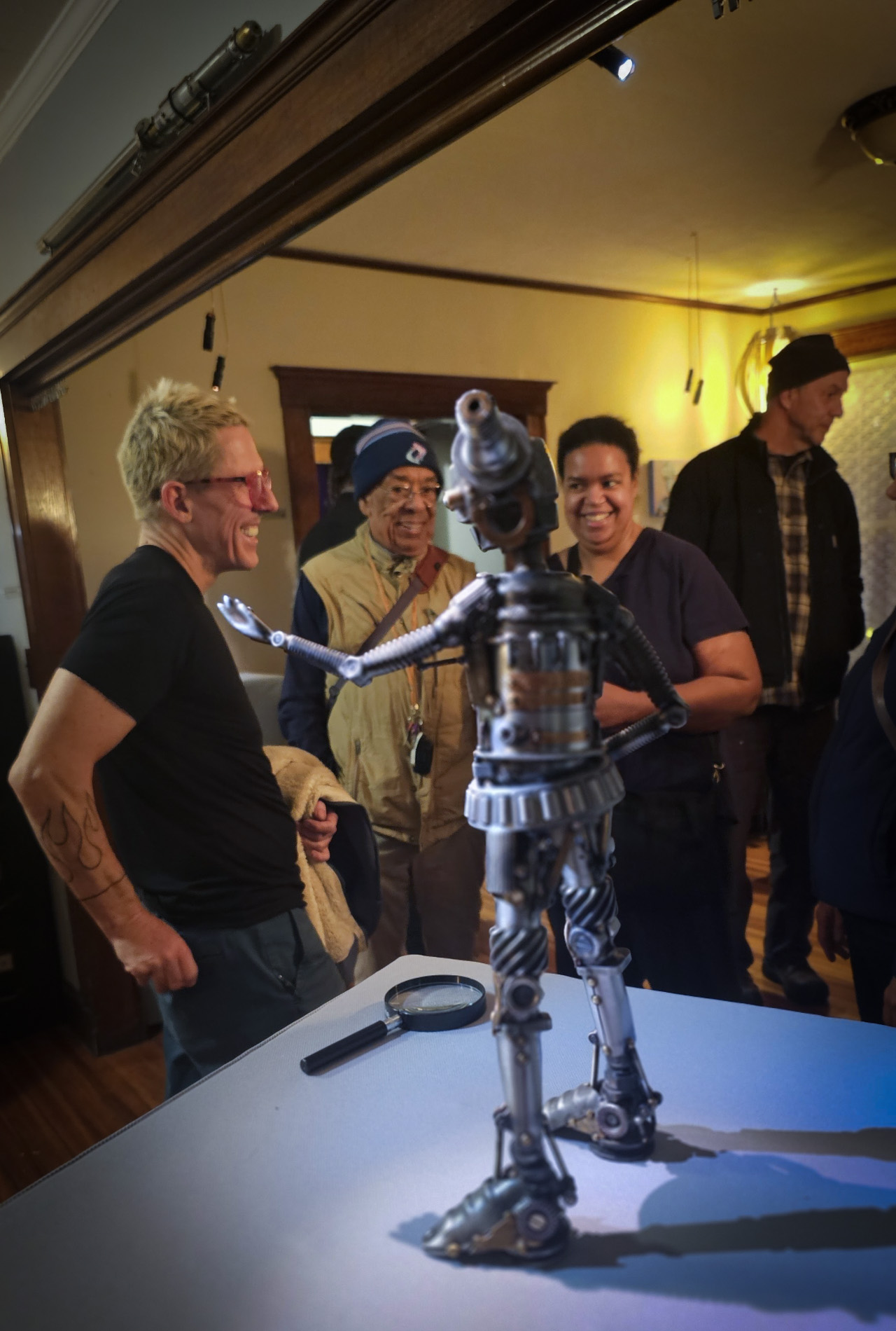 a robot sculpture is being admired by people at an art show. a magnifier lies on the pedestal next to the sculpture