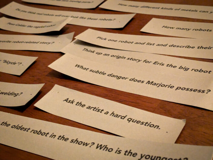 lots of slips of paper on a table, the foremost one being "ask the artist a hard question"