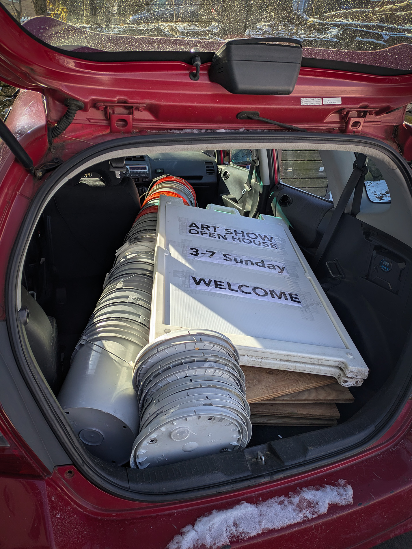 a long stack of bucket and a sandwich board sign advertising an open house and art show fills the inside of a compact car