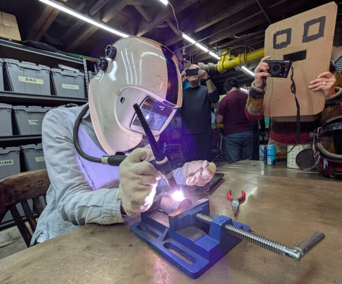 A welder tries her hand at TIG welding around the cirumference of a tube while others observe